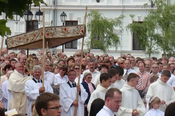 Procesja religijna z monstrancją, tłum ludzi w białych szatach, budynek w tle.