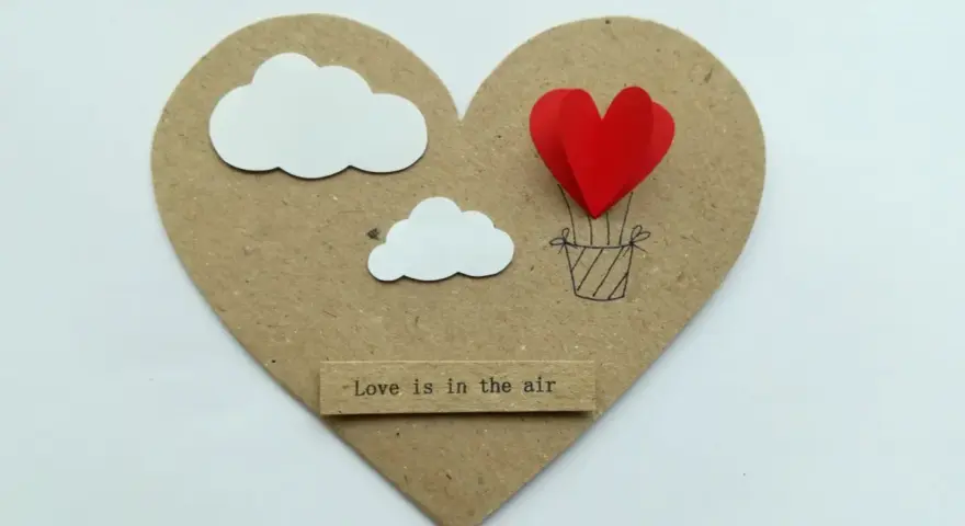 Serce z balonów w kształcie serca, unoszące się wśród chmur. Napis "Love is in the air".