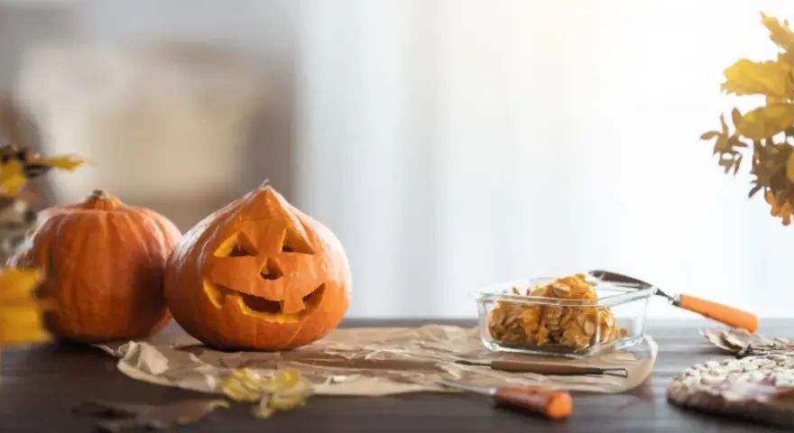 Wesoła dynia z wyciętą buzią i jej koleżanka czekają na Halloween. W szklanym naczyniu pestki dyni, gotowe na nowe pomysły na dynie.