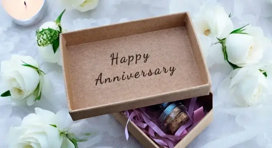 Otwarta szkatułka z napisem "Happy Anniversary" i małym prezentem w środku, otoczona białymi różami i płonącą świecą. Idealna na rocznice ślubu.