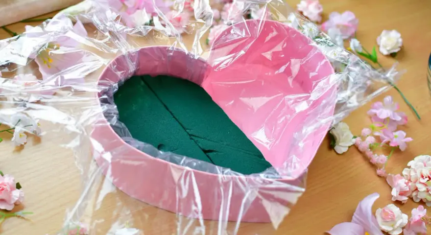 Jak zrobić flower box z winem? Różowe serce wyłożone folią i gąbką florystyczną, otoczone sztucznymi kwiatami.