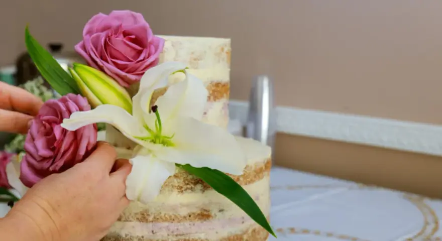 Dwie ręce dekorują dwupoziomowy tort żywymi margaretkami, różami i lilią.