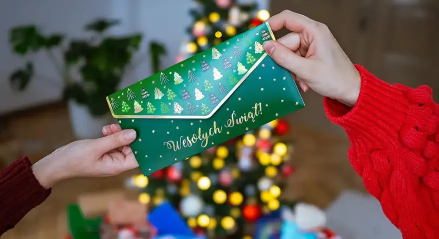 Jak zapakować voucher na prezent? Zielona koperta z choinkami i napisem "Wesołych Świąt!" jest przekazywana w świątecznej atmosferze.