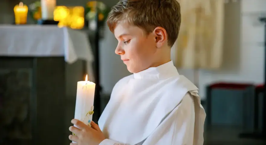 Chłopiec w bieli trzyma zapaloną świecę podczas uroczystości chrztu świętego.