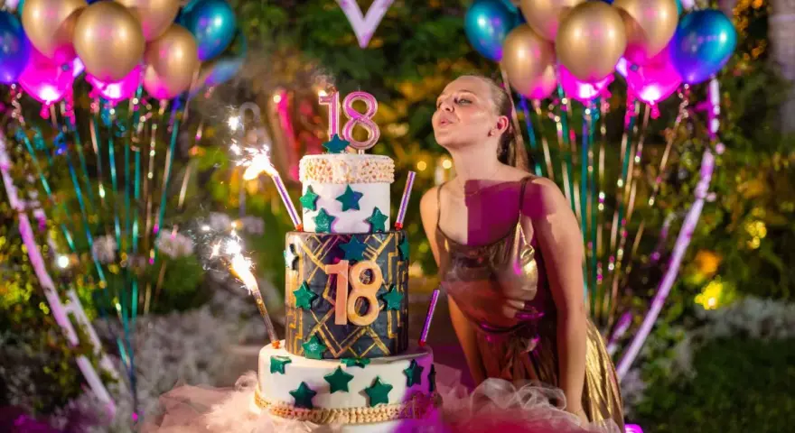Dziewczyna zdmuchuje świeczki na torcie z okazji 18. urodzin. Balony i fajerwerki podkreślają uroczystość.