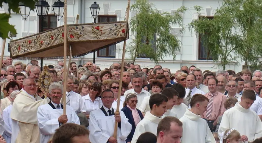Procesja religijna z monstrancją, tłum ludzi w białych szatach, budynek w tle.