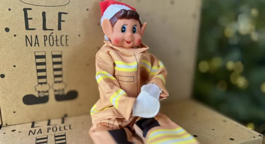 Uroczy elf na półce w stroju strażaka siedzi na kartonie z rysunkami elfich nóg.