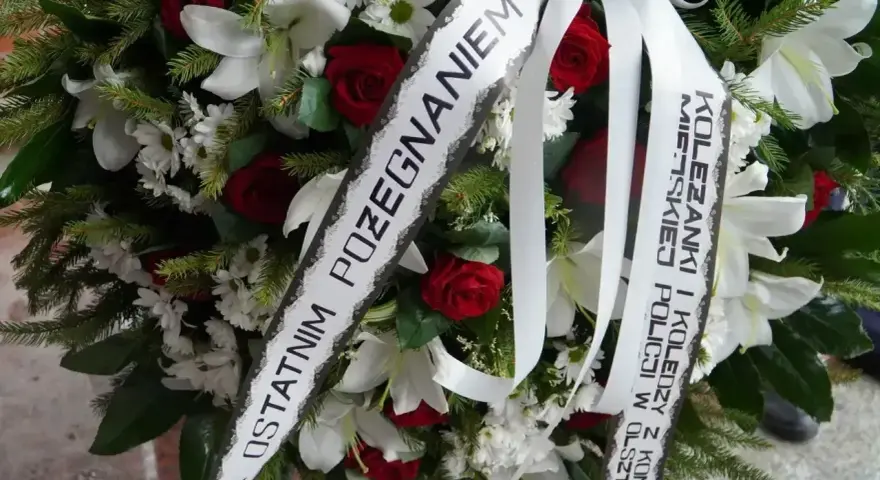Wieniec pogrzebowy z białymi liliami i czerwonymi różami. Napis na szarfie: "Z ostatnim pożegnaniem od koleżanki i kolegów z Komendy Miejskiej Policji w Olsztynie".