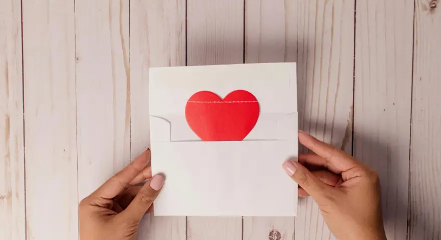 Dłonie trzymają białą kopertę z czerwonym sercem. To symboliczne podziękowania za spotkanie, pełne ciepłych uczuć.