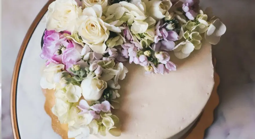 Elegancki tort z kremową polewą, ozdobiony bukietem żywych goździków, róż i hortensji w pastelowych kolorach.