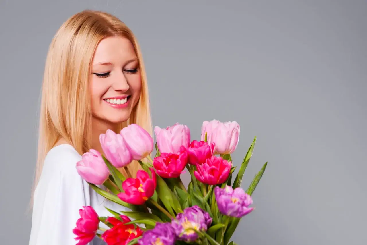Uśmiechnięta kobieta z blond włosami trzyma bukiet kolorowych tulipanów.