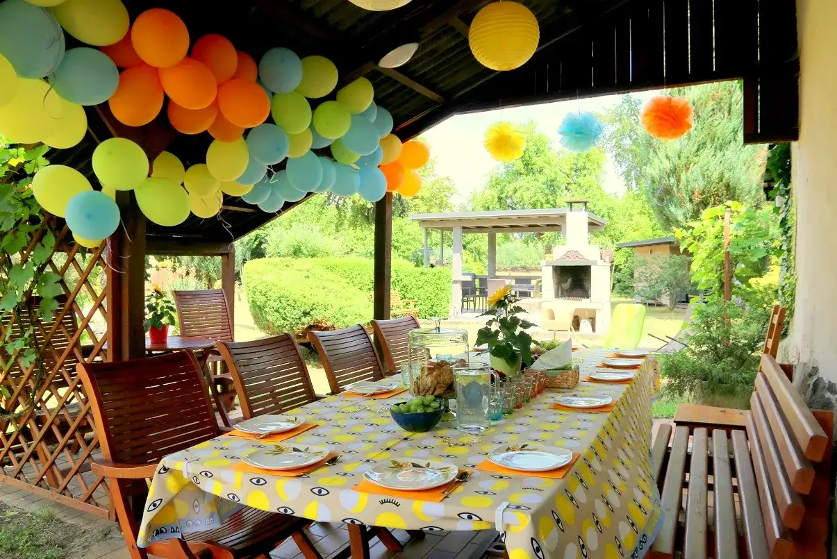Kolorowe balony i girlandy zapowiadają radosne urodziny na dworze. Stół nakryty do przyjęcia, a w tle grill i zielony ogród.
